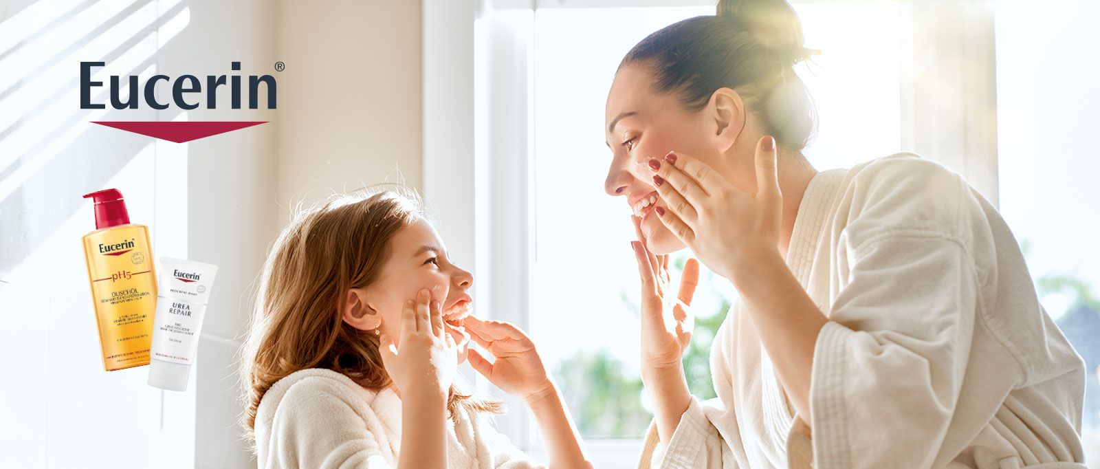 Lächelnde Mutter und Tochter in weißen Bademänteln tragen gemeinsam Hautcreme auf. Im unscharfen Hintergrund sind Eucerin-Hautpflegeprodukte zu sehen.