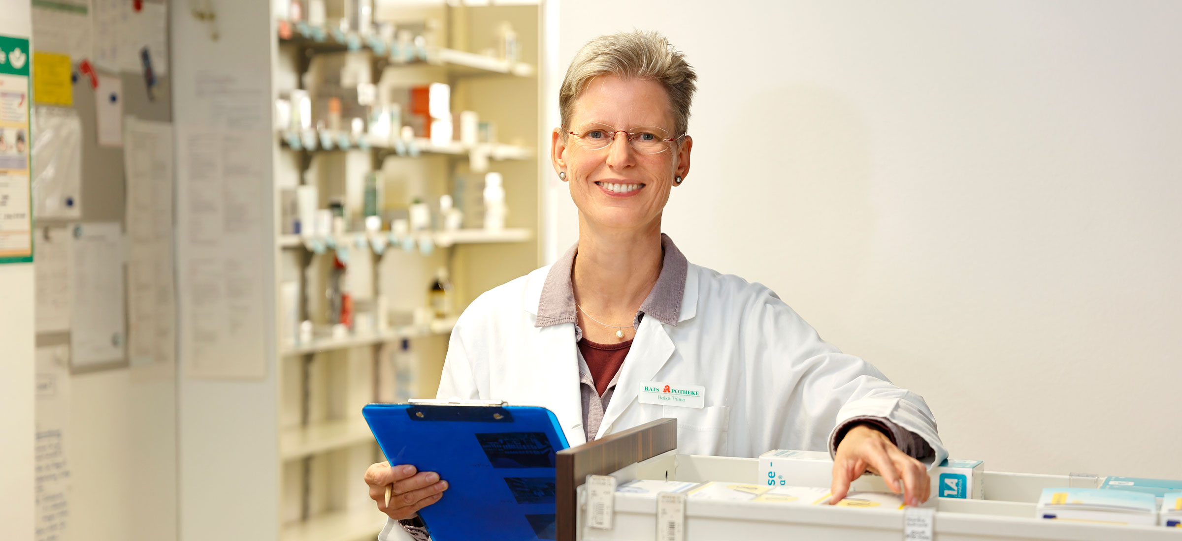 Freundliche Apothekerin im weißen Kittel lächelt mit einem Klemmbrett in der Hand an der Verkaufstheke.