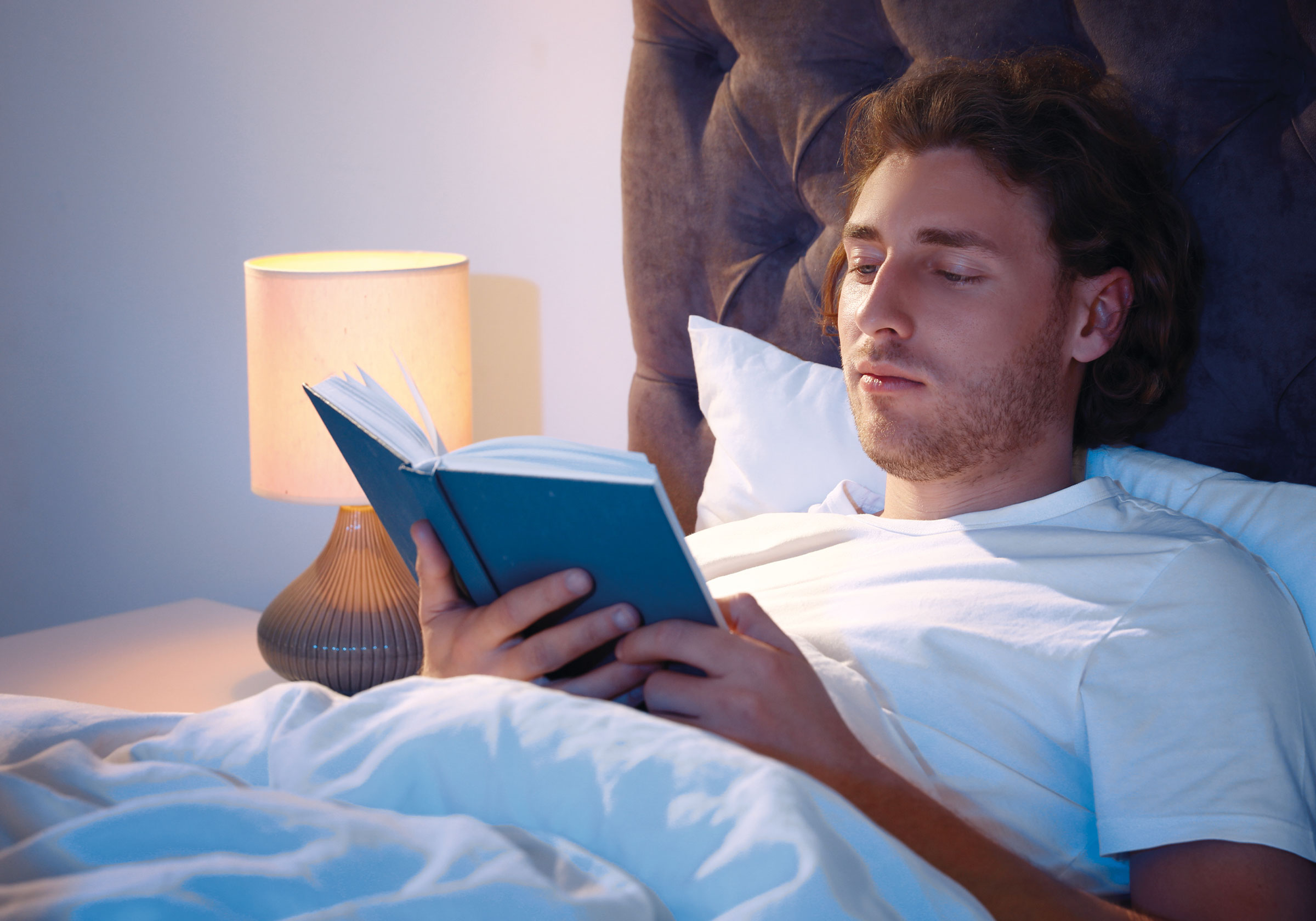 Ein Mann liest im Bett ein Buch, symbolisiert Entspannung und Schlaf.